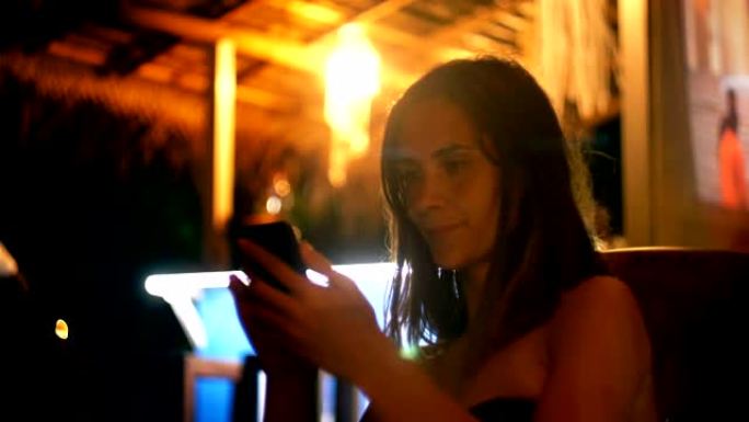 快乐放松的年轻白人妇女在度假时使用智能手机娱乐应用程序在大气的夜间休息室咖啡馆。