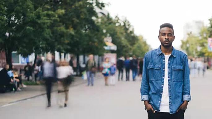 孤独的非洲裔美国男生的延时性学生站在大城市的步行街上，双手插在口袋里，当绕道者呼啸而过时，看着相机。
