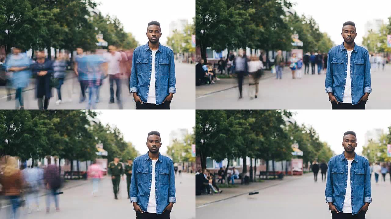 孤独的非洲裔美国男生的延时性学生站在大城市的步行街上，双手插在口袋里，当绕道者呼啸而过时，看着相机。