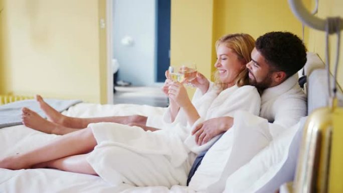 快乐的混血年轻成年夫妇躺在酒店房间的床上，穿着浴袍，手里拿着一杯酒，特写
