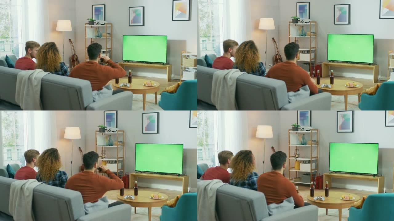 三个朋友坐在家里的沙发上，一边吃零食和喝饮料，一边看绿色色度键屏幕电视。年轻人在家里玩得开心。