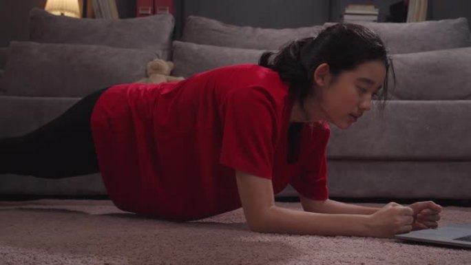 迷人的亚洲少年在家里客厅的笔记本电脑上观看和做木板运动，以增强核心力量，在线瑜伽直播课。她在应用程序