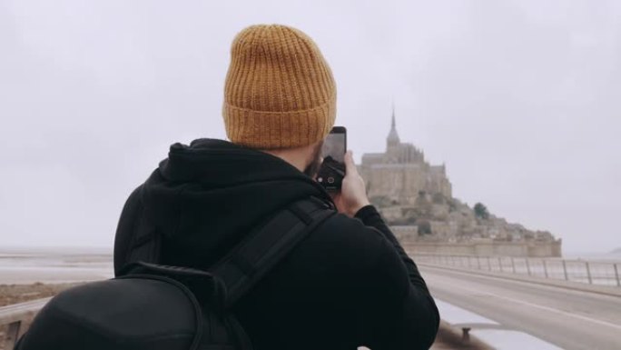 后视图快乐兴奋的旅游男子拍摄诺曼底史诗圣米歇尔山城堡的智能手机视频慢动作。