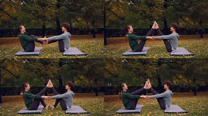 快乐的年轻女性正在做双人瑜伽，开心地坐在公园草地上的垫子上大笑。美丽的秋天自然草坪和树木是背景。