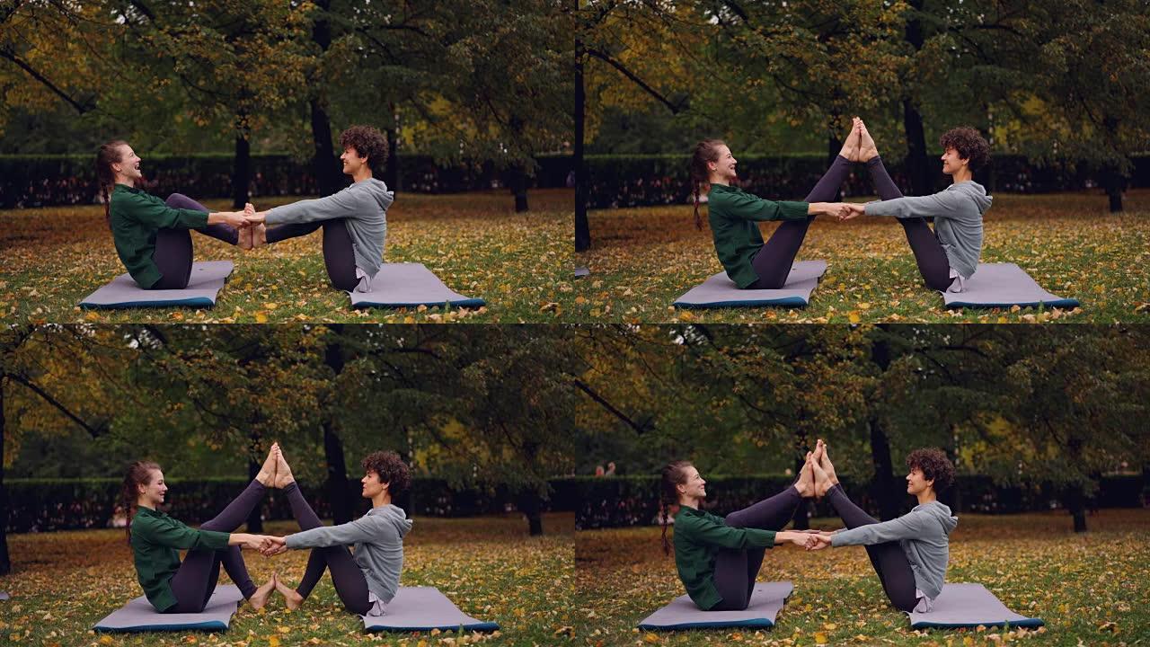快乐的年轻女性正在做双人瑜伽，开心地坐在公园草地上的垫子上大笑。美丽的秋天自然草坪和树木是背景。