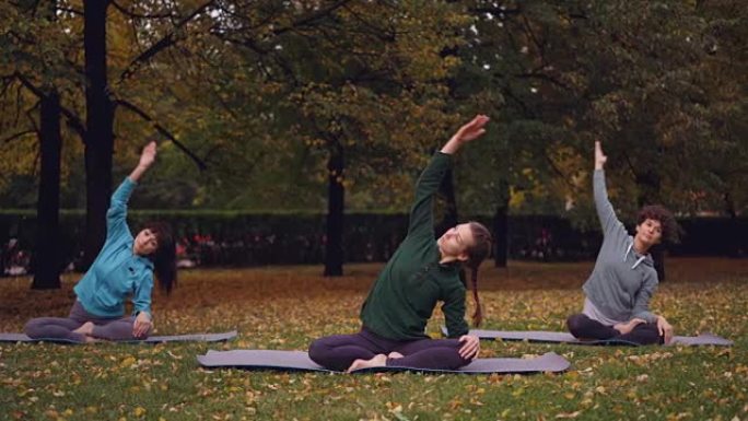 在公园的露天练习中，女瑜伽教练正在展示伸展运动，然后在荷花姿势放松，闭着眼睛坐在垫子上。