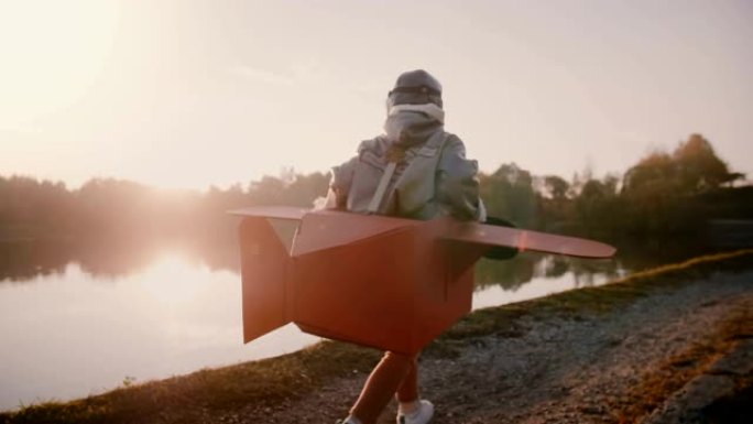 摄像机跟随快乐的欧洲男孩在日落森林河上奔跑，穿着有趣的服装慢动作扮演飞机飞行员。