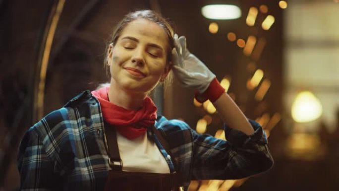 年轻美丽的赋权女人摩擦额头，对着镜头轻轻微笑。真正的制造商在金属车间穿着工作服。火花在背景上飞舞。