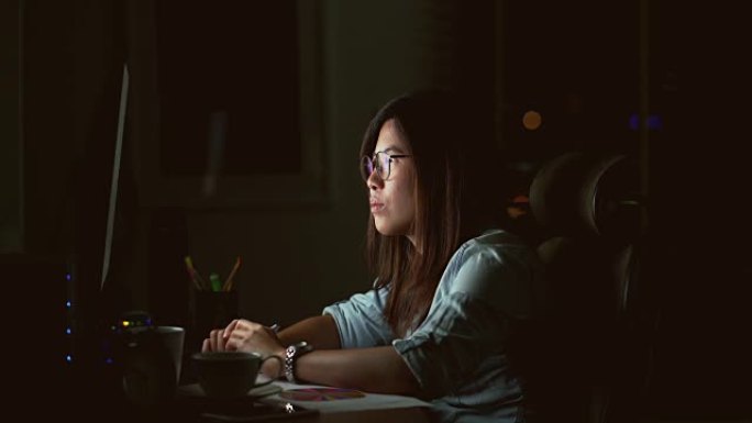 4k镜头的场景有吸引力的亚洲女性工作到很晚，在黑暗中工作场所的计算机显示器桌面前的桌子上认真思考，工