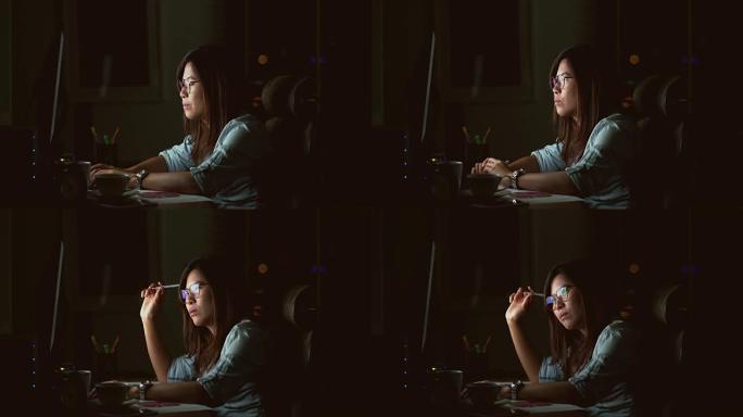 4k镜头的场景有吸引力的亚洲女性工作到很晚，在黑暗中工作场所的计算机显示器桌面前的桌子上认真思考，工