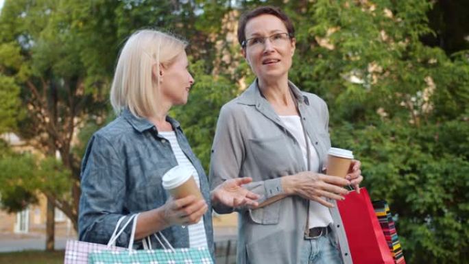 快乐的女人带着明亮的购物袋和咖啡在公园散步聊天