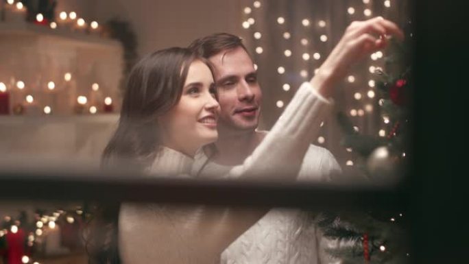 一对年轻夫妇正在家里装饰圣诞树，他们快乐地庆祝圣诞节。通过房子的窗户从外面看摄像机。