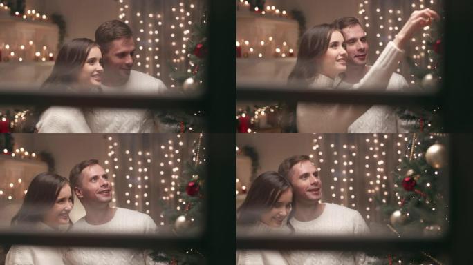 一对年轻夫妇正在家里装饰圣诞树，他们快乐地庆祝圣诞节。通过房子的窗户从外面看摄像机。