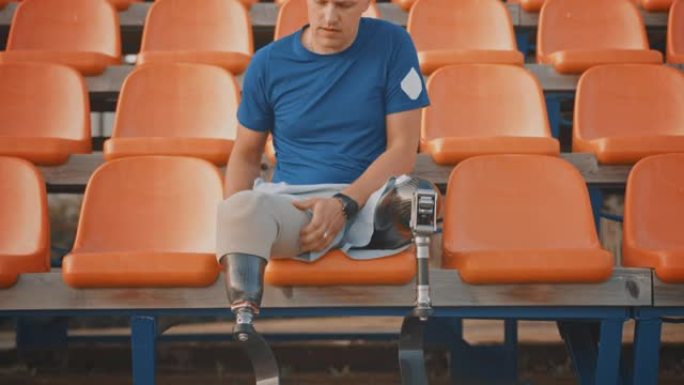 带有假肢跑步刀片的运动残疾人健身男子正在一个阳光明媚的下午准备在室外体育场进行训练。截肢者正坐着，固