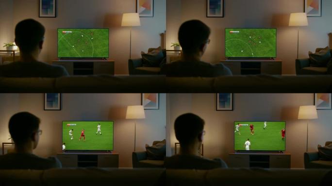 戴眼镜的年轻人正坐在沙发上看足球比赛的电视。现在是晚上，家里的房间有工作灯。