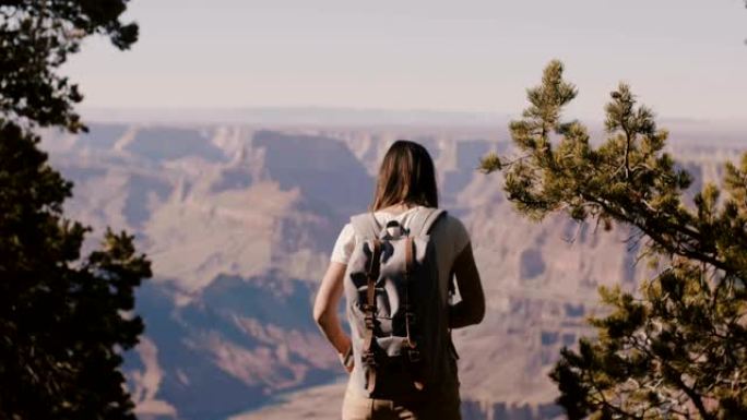 美丽的年轻快乐的旅游女人背着背包看着史诗般的大峡谷山脉，走回相机。