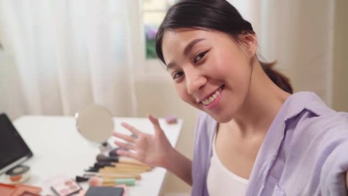 快乐美丽的年轻亚洲女性使用智能手机自拍在线直播到社交网络。美容博主展示美容化妆品坐在平板电脑摄像头前