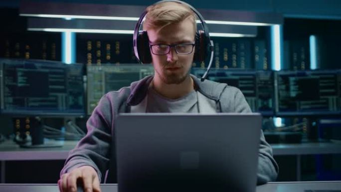 软件开发人员/黑客/游戏玩家的肖像，戴着眼镜和耳机坐在办公桌前，在笔记本电脑上工作/玩。在背景黑暗的
