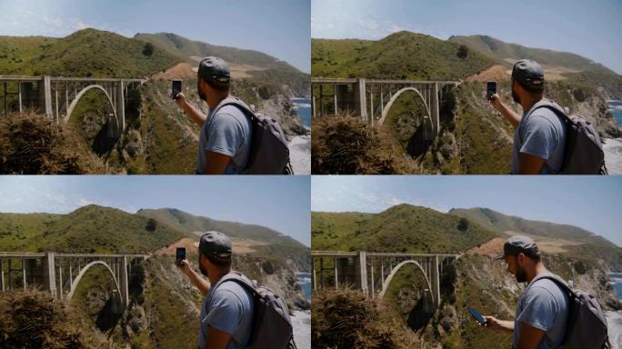 年轻快乐兴奋的背包旅游男子拍摄了加州大苏尔标志性比克斯比溪桥的智能手机照片。