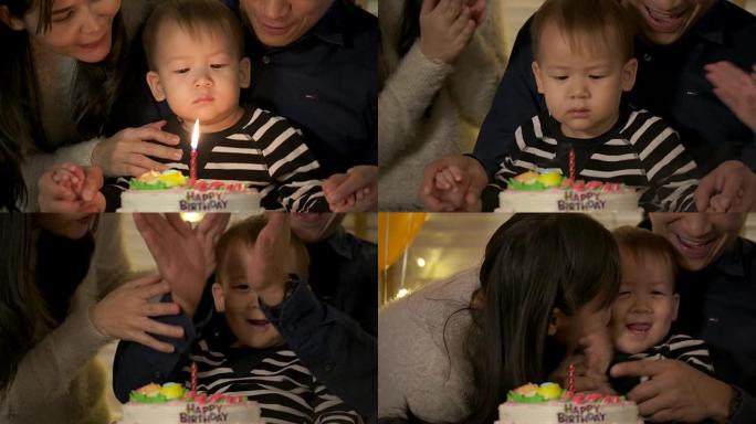 一个可爱的小男孩坐在桌子旁，在生日蛋糕上吹蜡烛，而他的家人站在后面，为他唱歌。慢动作。
