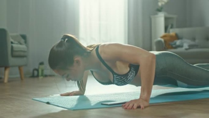 特写镜头，一个美丽的健身女孩在运动上衣上做俯卧撑练习，同时在手机上使用秒表。她正在家里的客厅里训练，