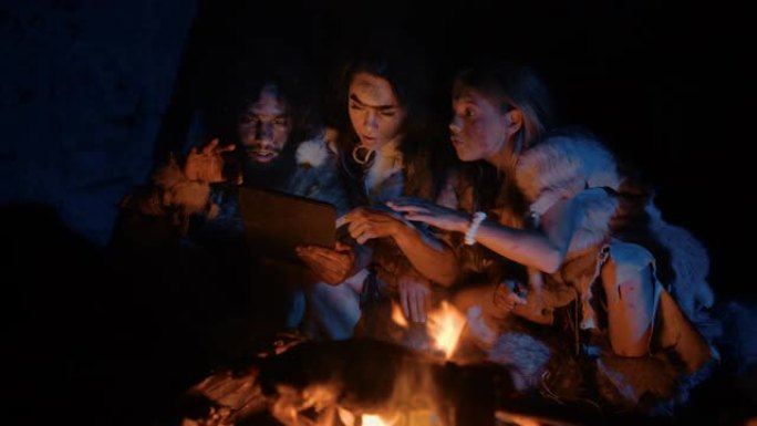 穿着兽皮的史前原始狩猎采集者部落在夜间在洞穴中使用数字平板电脑。尼安德特人或智人家庭浏览互联网，观看