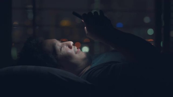 亚洲男子在夜间与bokeh light city一起在床上使用智能手机
