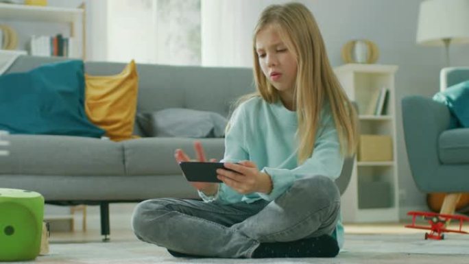 聪明可爱的女孩坐在家里的地毯上玩智能手机上的视频游戏，在水平景观模式下拿着和使用手机。孩子在阳光明媚