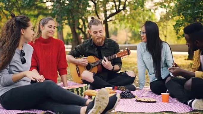 年轻人吉他手的慢动作在户外野餐时唱歌和弹吉他，而他的朋友们则在听，笑和拍手。