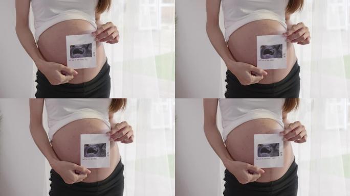 一名孕妇将手放在腹部，并在家里的室内拿着超声波图像。