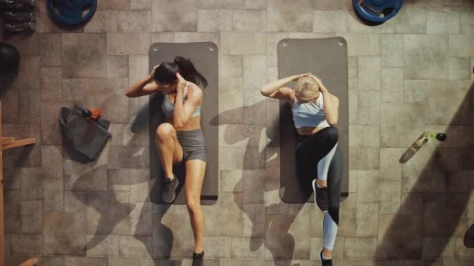 两名专业女性健美运动员躺在硬核健身房的瑜伽垫上做自行车仰卧起坐的俯视图。肌肉和运动美丽的女孩肌肉，力