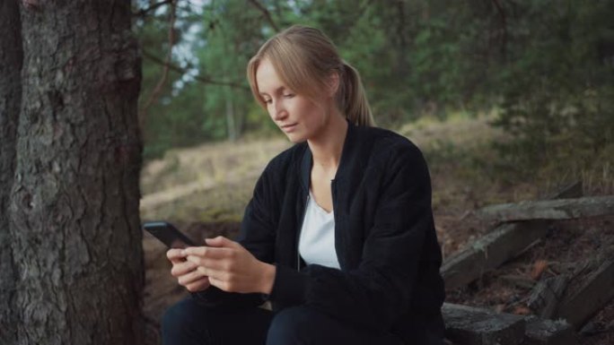 一个年轻美丽的金发女人在浪漫的自然氛围中的肖像。女孩穿着黑色衣服，正在使用她的智能手机。她在网上浏览