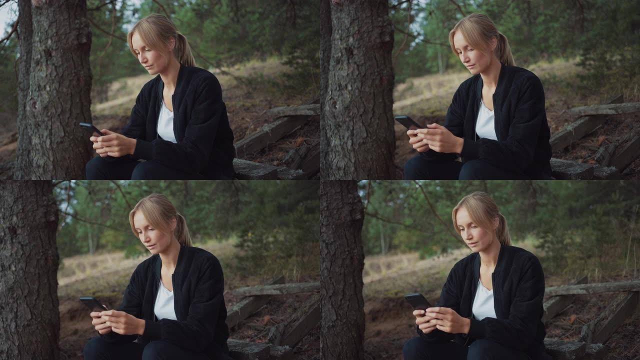 一个年轻美丽的金发女人在浪漫的自然氛围中的肖像。女孩穿着黑色衣服，正在使用她的智能手机。她在网上浏览