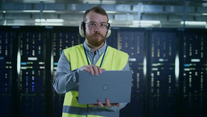 戴着眼镜和耳机的大胡子IT专家，穿着高可见度背心，正在服务器机架旁的数据中心的笔记本电脑上工作。他合