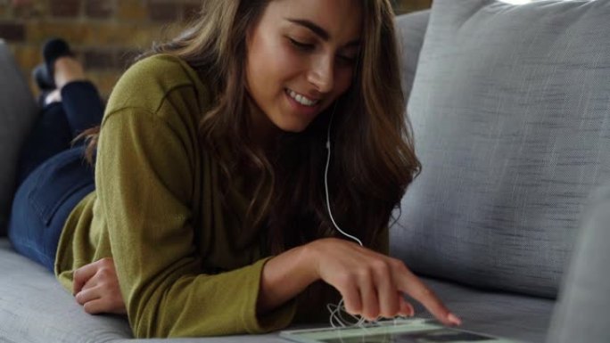 女年轻女子躺在沙发上看着平板电脑上的照片，同时戴着耳机听音乐