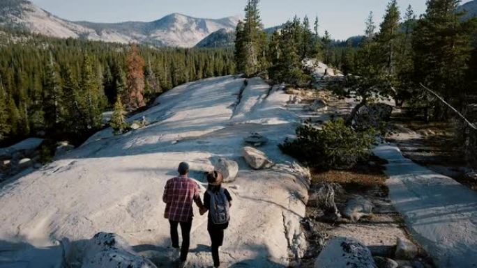 无人机摄像机跟随年轻浪漫的情侣一起沿着约塞米蒂国家公园森林的惊人白岩徒步旅行。