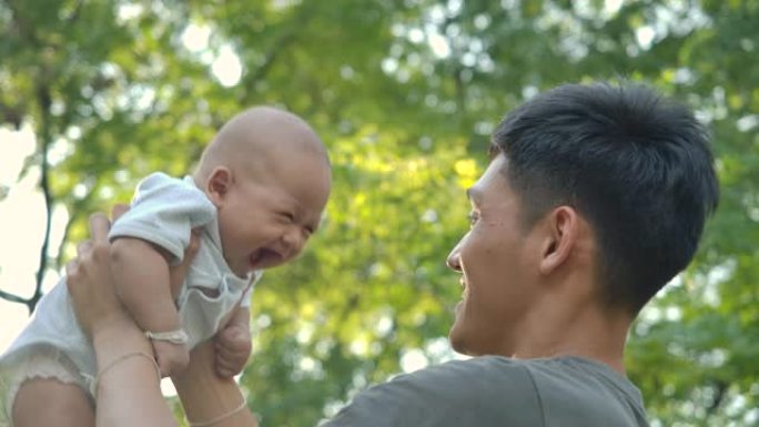 父亲在公园和婴儿玩耍。年轻的亚洲父亲带着快乐的情感在公园或花园高高举起他的宝贝儿子。慢动作。