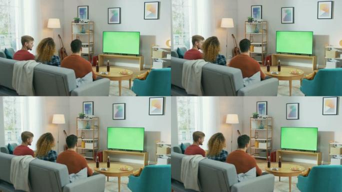 三个朋友坐在家里的沙发上，一边吃零食和喝饮料，一边看绿色色度键屏幕电视。年轻人在家里玩得开心。