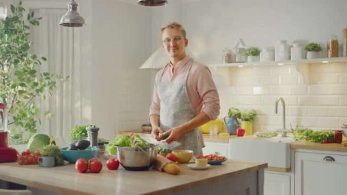 穿着粉红色衬衫和围裙的英俊男子正在现代阳光明媚的厨房里做一顿健康的有机沙拉。戴眼镜的潮人对着镜头微笑