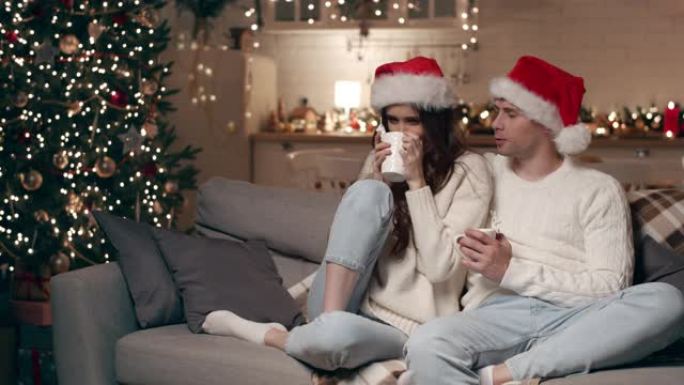 一对相爱的夫妇坐在家里的沙发上，圣诞节晚上，他们喝白色杯子里的热饮。