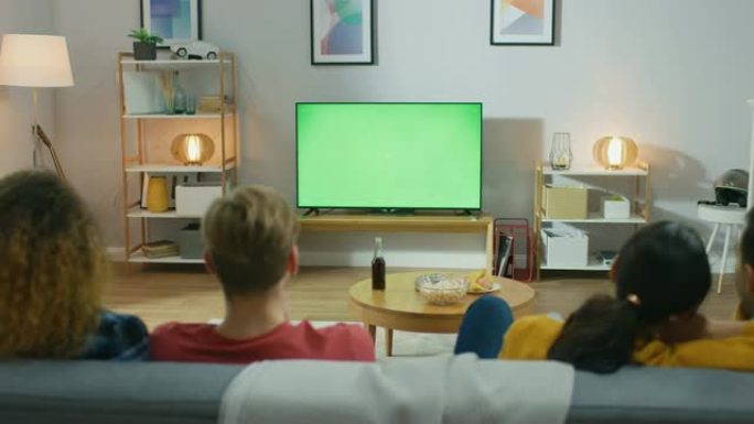 在家里，一群不同的朋友在绿色色度键屏幕电视上观看喜剧，坐在客厅里吃零食和喝饮料。年轻人表示关切和困惑