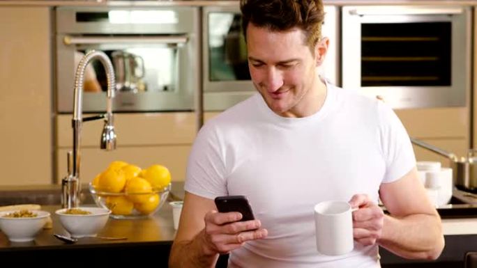 一个男人在厨房里吃早餐，在杯子里喝茶或牛奶时，用电话发送消息或打电话，并微笑。