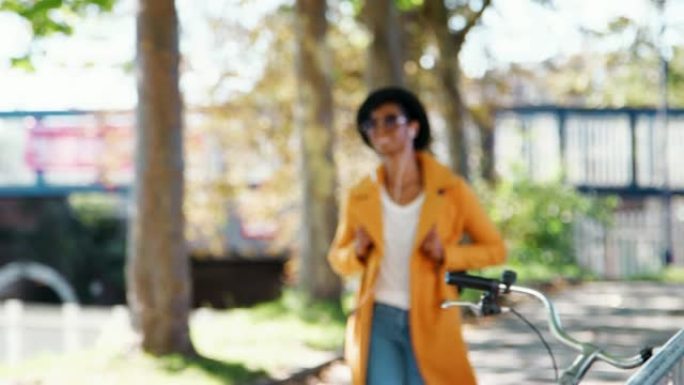 时尚的年轻黑人女性戴着帽子、太阳镜、蓝色牛仔裤和黄色豌豆大衣，在阳光明媚的日子里沿着树木覆盖的街道行