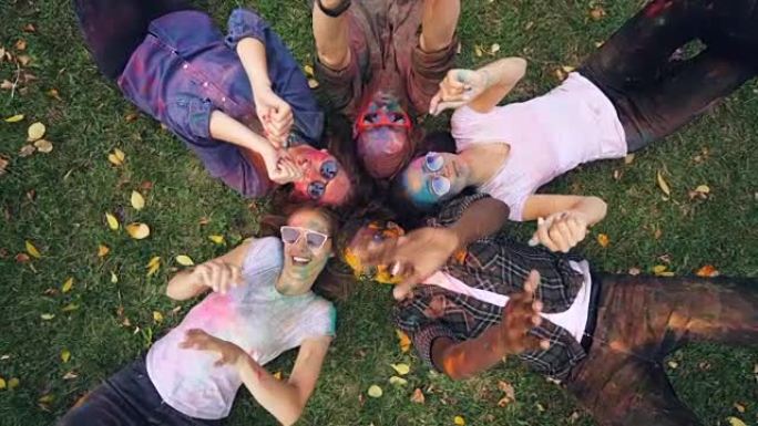 放松的人放大-多民族的人躺在草地上，带着彩色的脸和衣服，看着相机，微笑着移动的手。放松和派对概念。