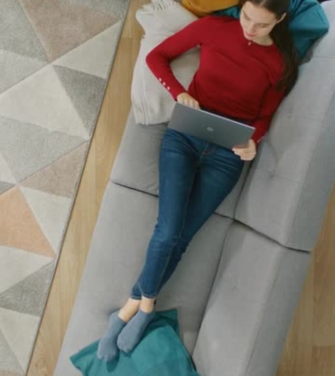 穿着红色上衣和蓝色牛仔裤的年轻女孩躺在沙发上，用笔记本电脑。她抬头微笑。放大俯视图。垂直屏幕方向9: