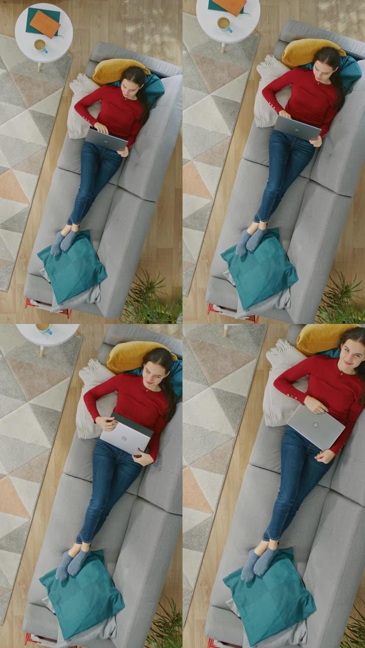 穿着红色上衣和蓝色牛仔裤的年轻女孩躺在沙发上，用笔记本电脑。她抬头微笑。放大俯视图。垂直屏幕方向9: