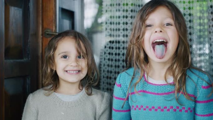 两个可爱的小女孩在棒棒糖后露出彩色舌头，并在屋外的相机中微笑的真实照片