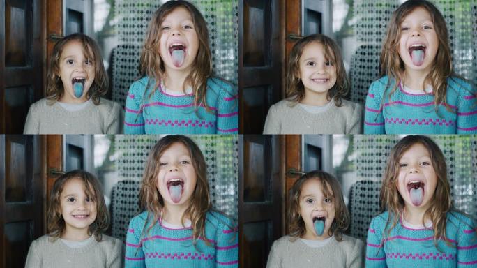 两个可爱的小女孩在棒棒糖后露出彩色舌头，并在屋外的相机中微笑的真实照片