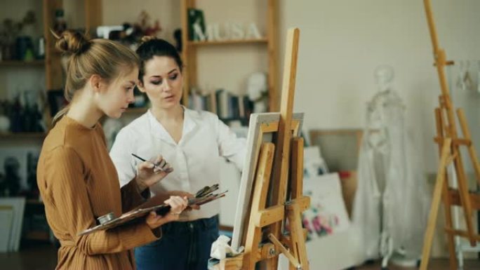 可爱的十几岁的女孩正在向工作室艺术学校的经验丰富的老师学习绘画，这些老师有艺术品和木制画架。艺术家正