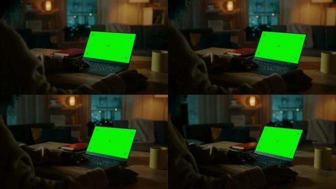 坐在办公桌前的美丽黑人妇女在带有绿色模拟屏幕的笔记本电脑上工作。深夜在她的客厅里，女孩使用笔记本电脑